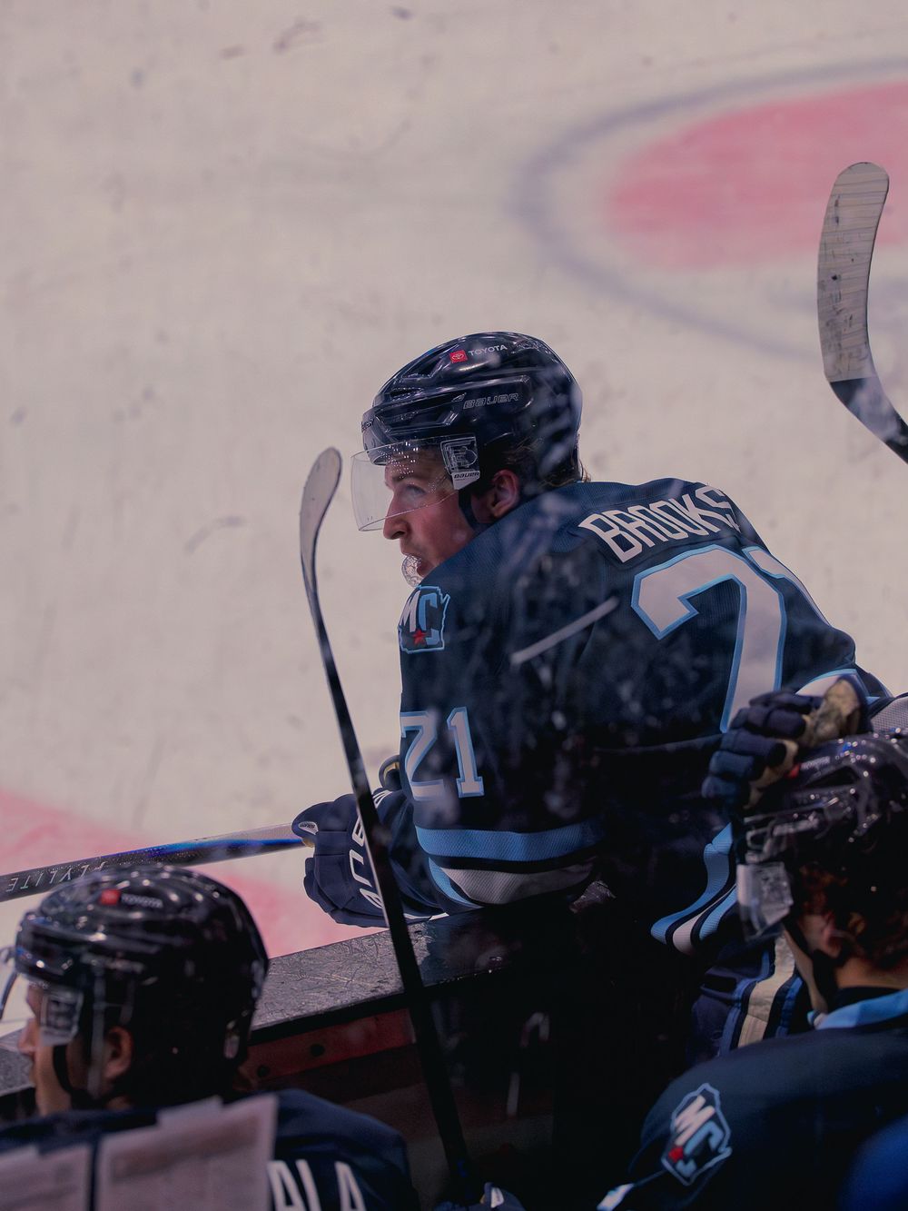 A Madison Capitols player number 21 leans over the boards at the bench, mouthguard out, watching the play with teammates alongside him.