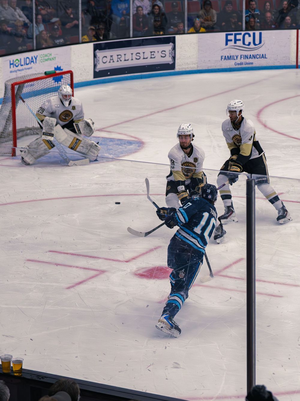 A Capitols player number 10 winds up a shot as two white Lumberjacks defenders and their white-clad goaltender number 29 guard the net.