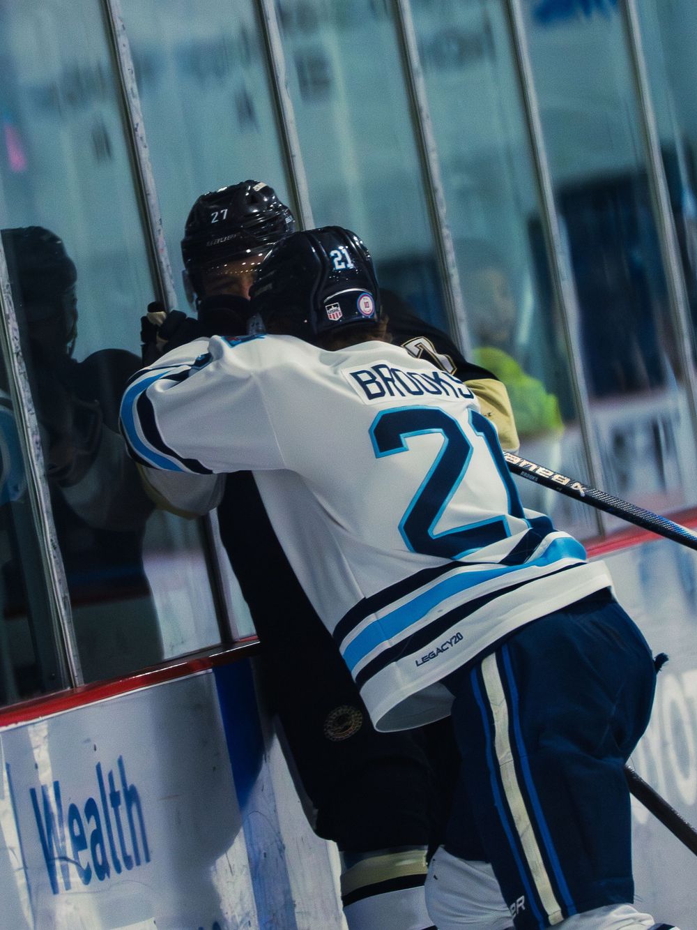 A Madison Capitols player number 21 pins a black-and-gold Lumberjacks player number 27 hard against the glass along the boards.