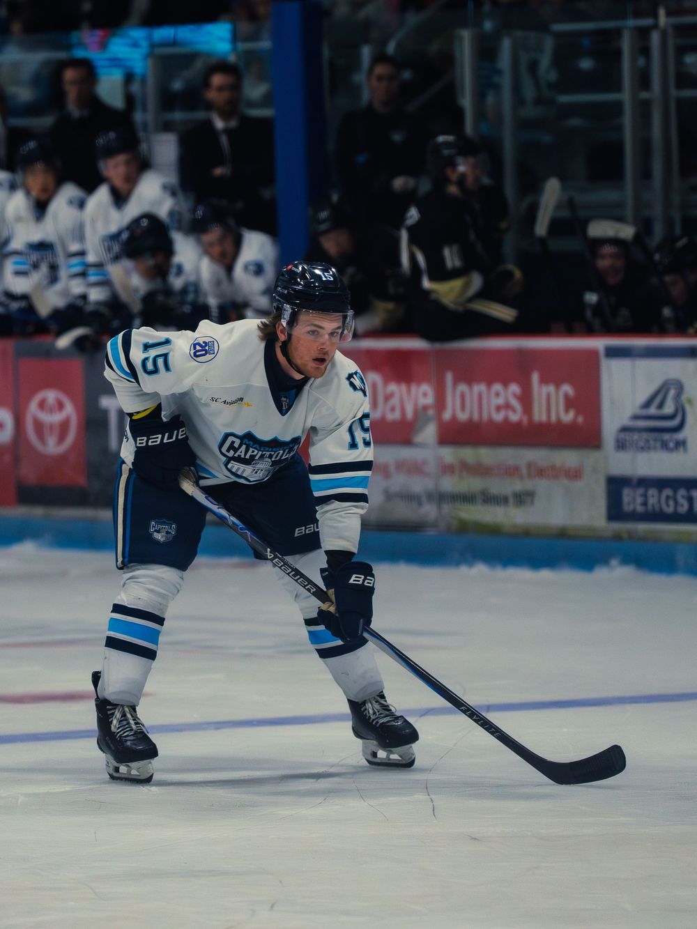 A Madison Capitols player in white number 15 crouches in a ready stance, stick on the ice, with the team bench and boards behind him.