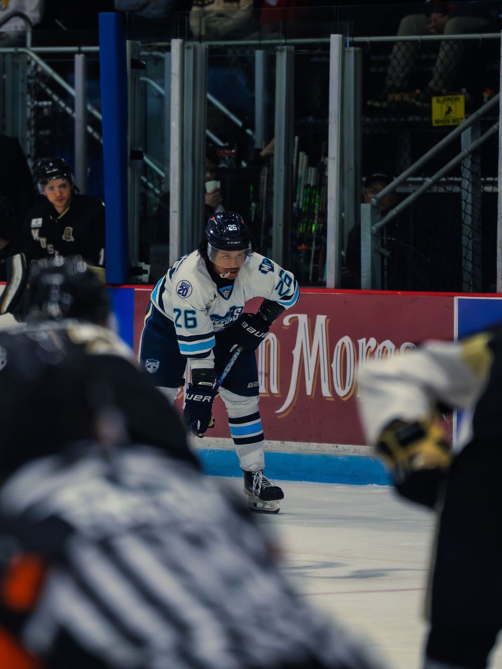 A Madison Capitols player in white number 26 crouches in a ready stance near the boards, framed between blurred players in the foreground.
