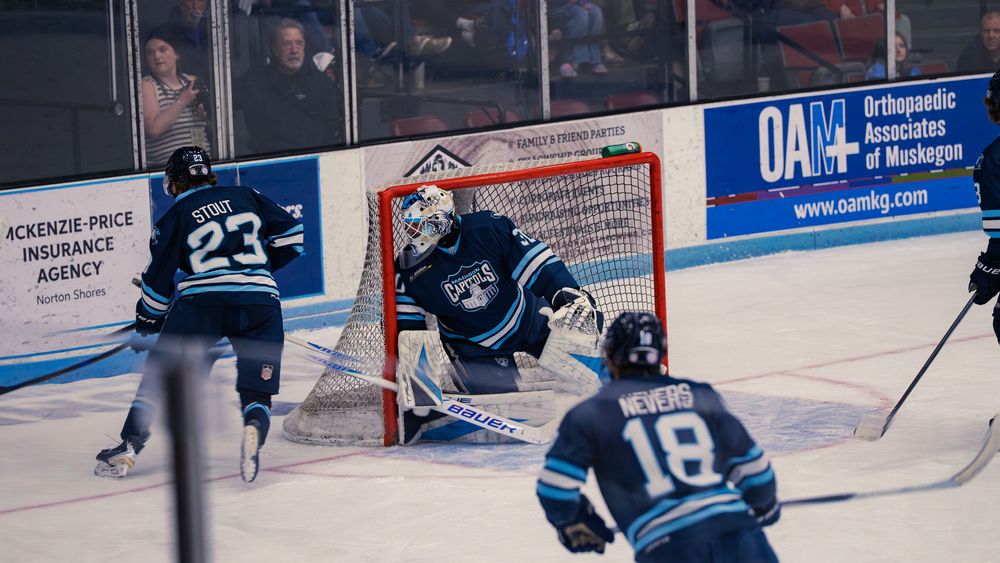 A Madison Capitols goaltender sets in his crease to face an incoming shot as two navy teammates skate in to defend around the net.