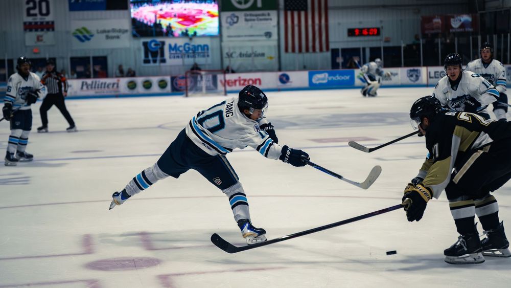 A Capitols player in a white number 10 jersey lunges to shoot the puck past a defending Lumberjacks player in black and gold on the ice.