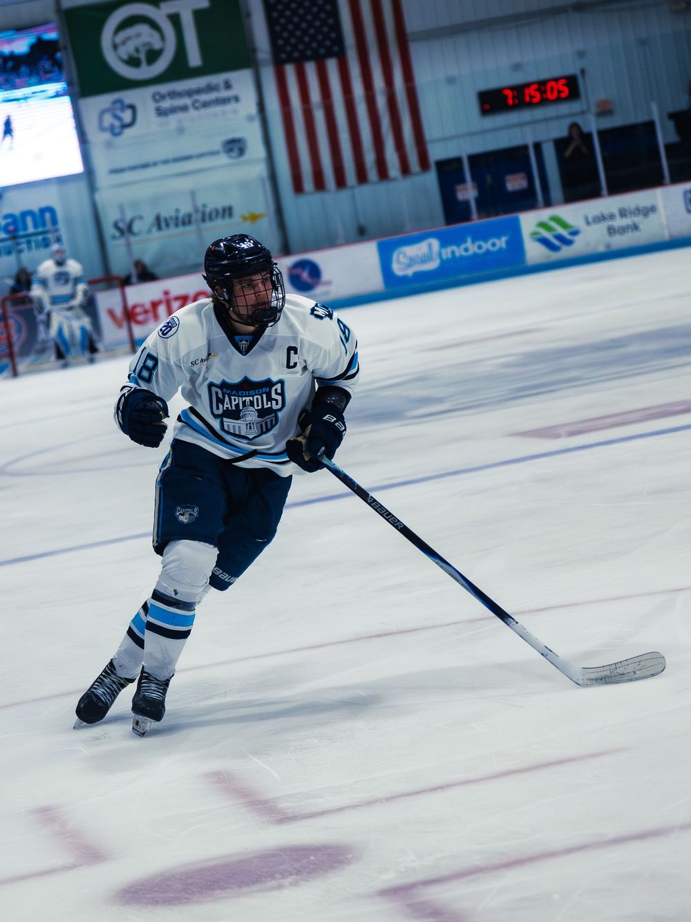 A Madison Capitols player in white number 18 with a captain's C skates up ice, stick down, the goaltender defending the net behind him.