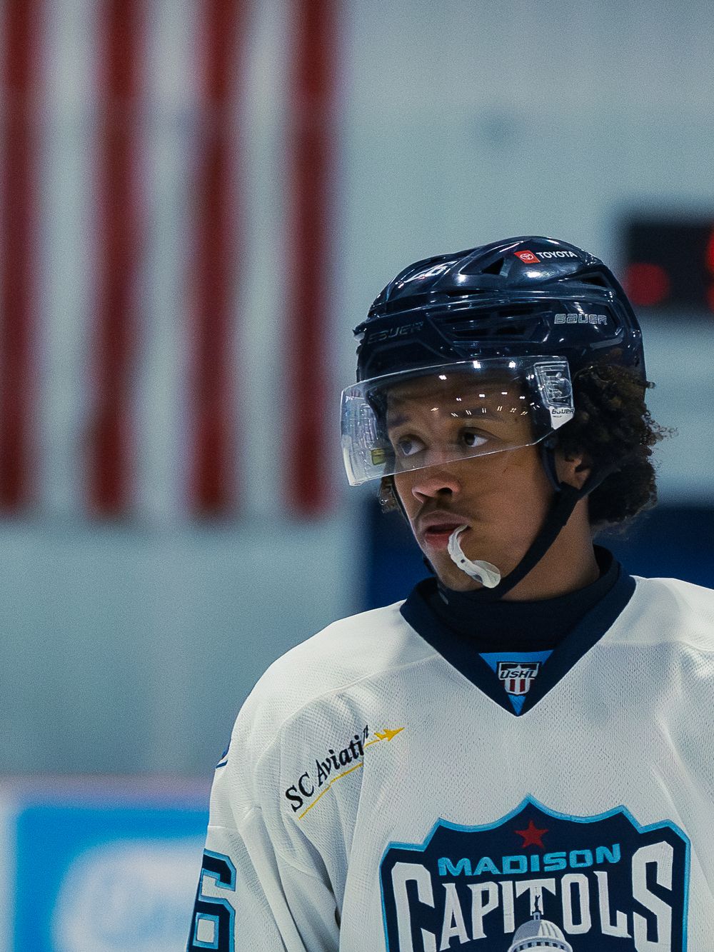 A close-up of a Madison Capitols player in a white jersey and navy helmet, mouthguard out, looking to the side, capitol-dome crest visible.