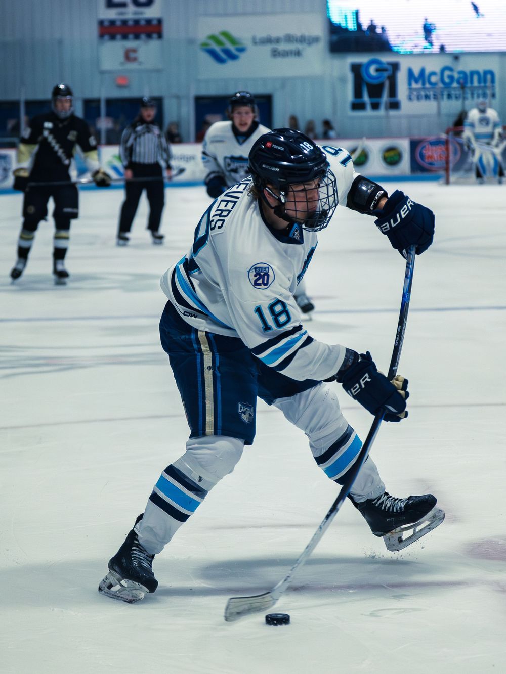 A Madison Capitols player in white number 18 winds up to shoot the puck as teammates, an opponent and a linesman look on in the background.