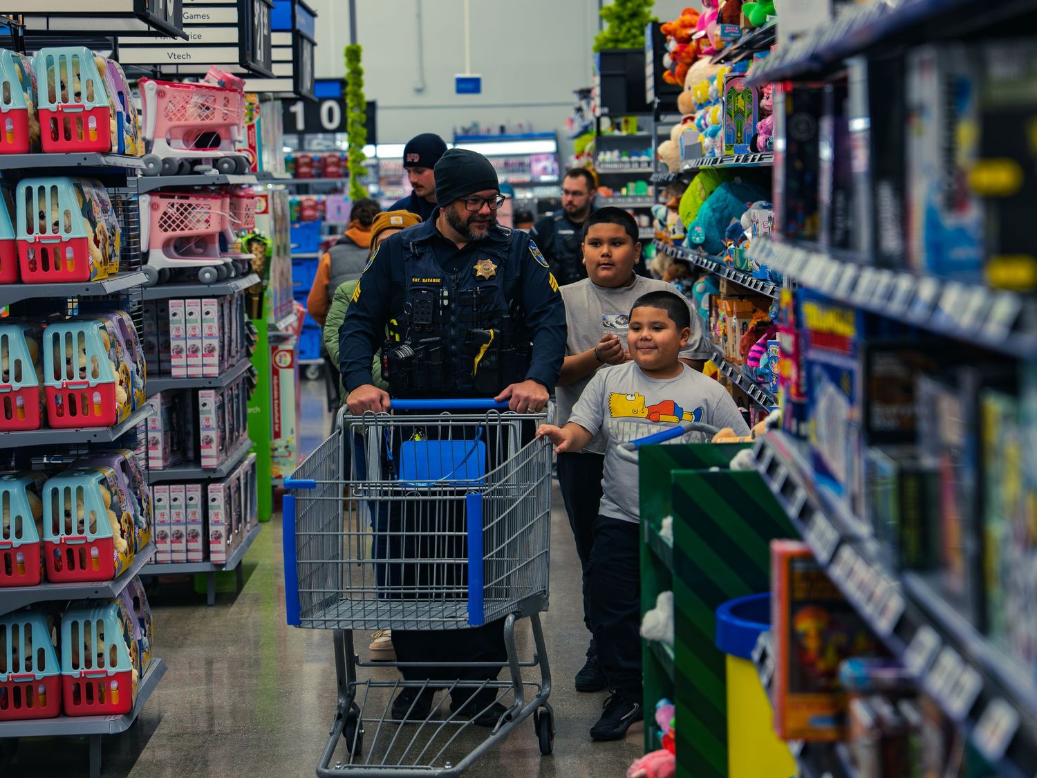 A police officer pushes a shopping cart down a toy aisle with two smiling children walking alongside as more officers shop behind them.