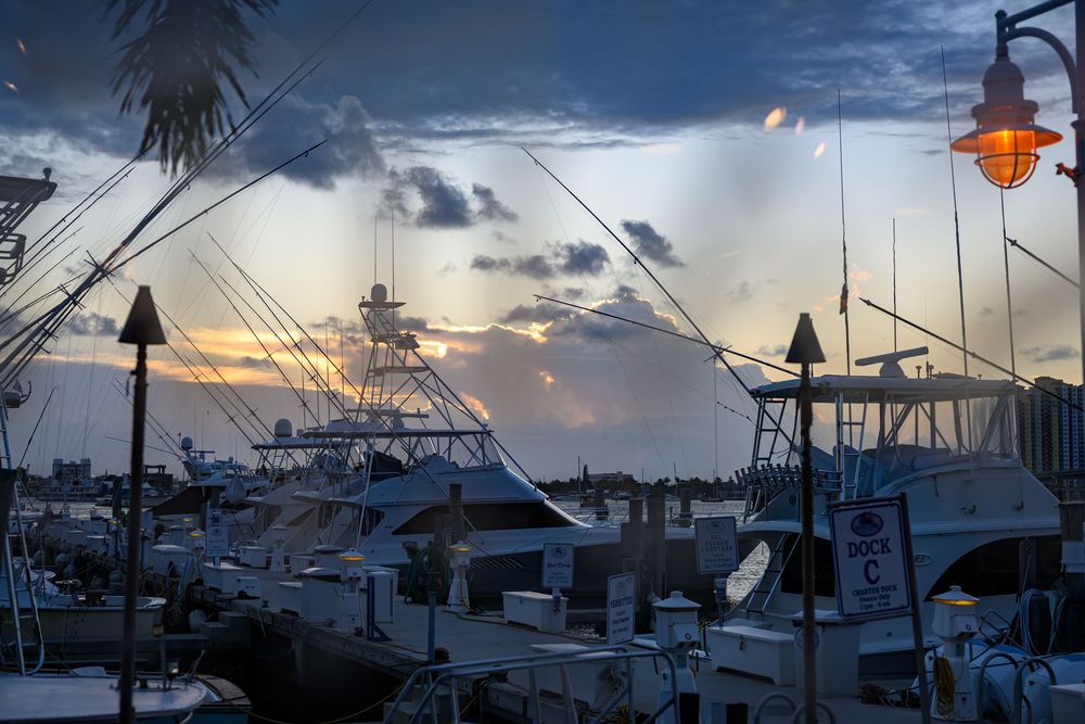 Boats docked at a marina at sunset with dramatic cloudy sky and glowing orange lamp.