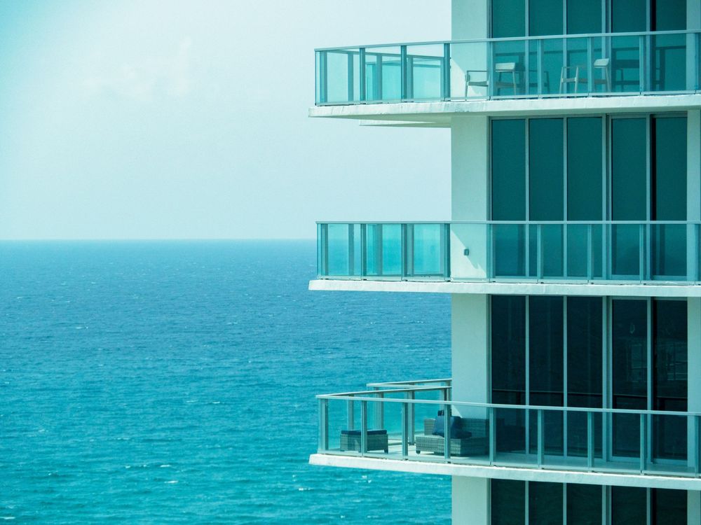 Modern oceanfront building balconies with glass railings overlooking turquoise blue sea.