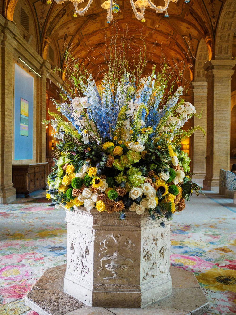 Elegant floral arrangement of blue, yellow, and white flowers in ornate stone urn inside historic hall.
