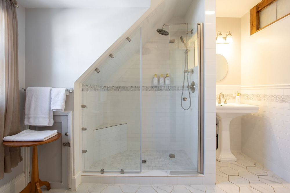 Modern glass-enclosed shower under a sloped ceiling with a pedestal sink and white tile bathroom.