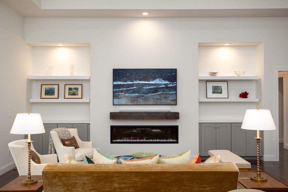 Bright living room with built-in shelves, electric fireplace, ocean artwork, and classic table lamps.