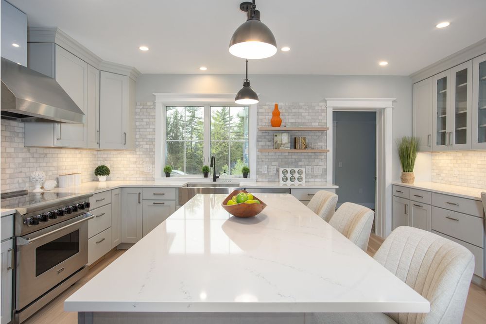Bright white kitchen with quartz island, gray shaker cabinets, herringbone backsplash, and stainless steel appliances.