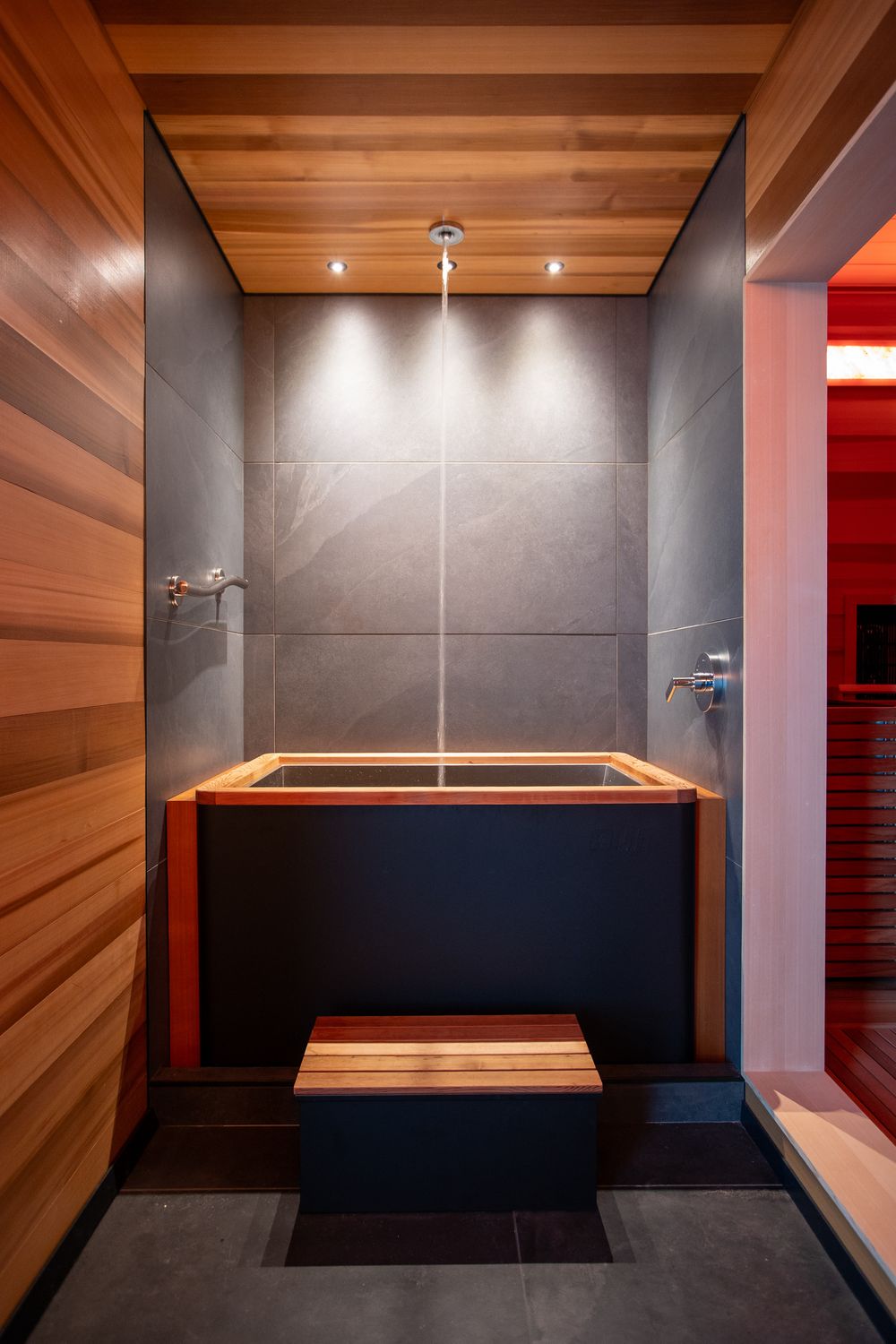 Japanese-style soaking tub with cedar accents, slate tile walls, overhead shower, and ambient lighting.