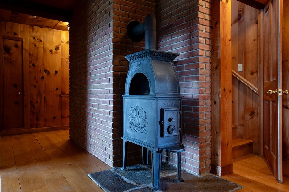 Ornate cast iron wood-burning stove on brick hearth with decorative floral design in rustic wood-paneled room.