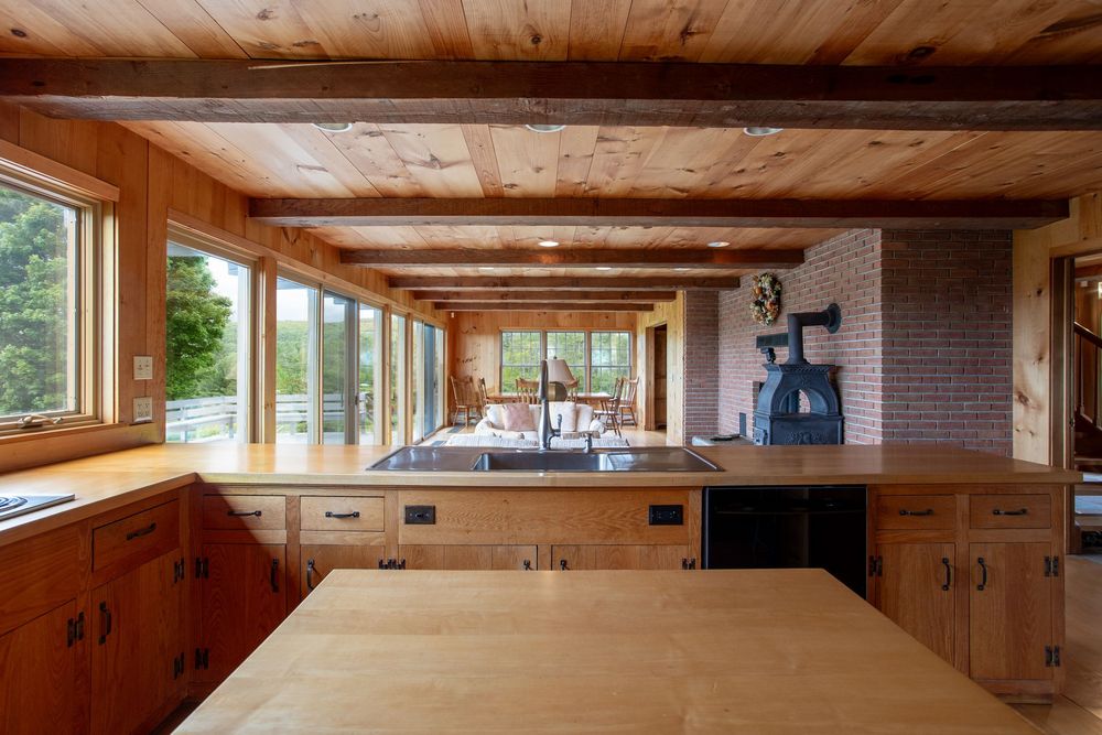 Rustic wood kitchen with beam ceiling, oak cabinets, center island, and large windows overlooking lush greenery.