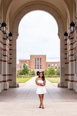 A young woman in a white dress poses under a grand stone arch on a university campus with a brick building behind her.