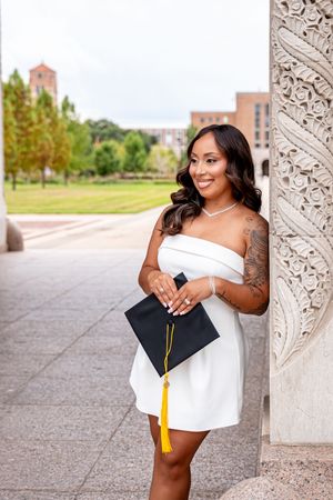 A smiling woman in a white dress holds a black graduation cap with yellow tassel, posing by an ornate column on campus.