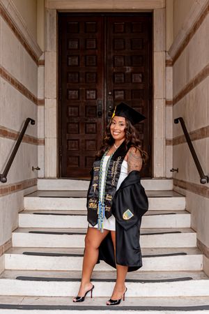 Smiling graduate in black cap and gown poses on steps before ornate wooden doors, wearing heels and honor stoles.