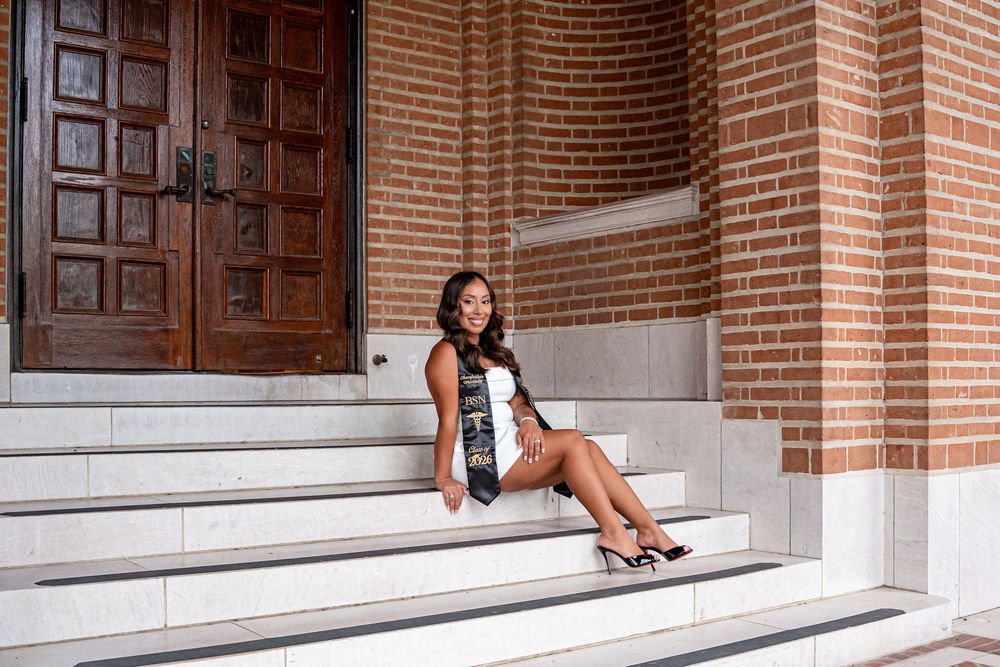 Young woman in graduation attire sitting on marble steps of a brick building with wooden doors.