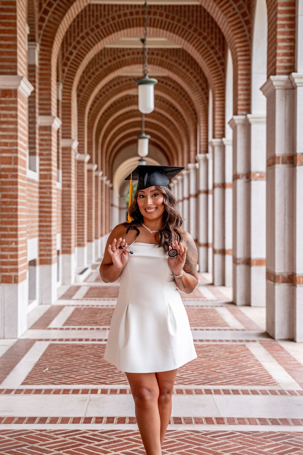 Smiling graduate in white dress and cap poses in brick archway corridor for graduation photos.