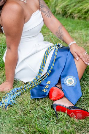 Graduate in white dress with BSN stole, honor cords, and red heels sitting on grass after nursing graduation ceremony.