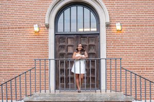 Young woman in white dress posing on brick building steps by ornate wooden door with arched window.