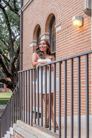 A smiling woman in a white dress leans on a black railing outside a red brick building with arched windows.