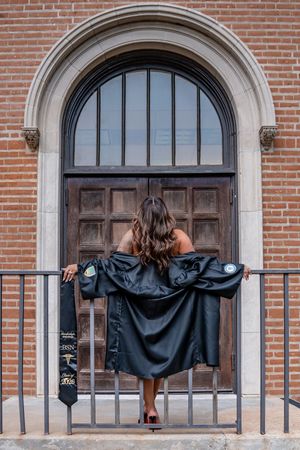 Graduate poses with black academic gown spread wide before ornate brick building arched doorway.
