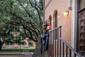Graduate in cap and gown posing on brick building steps under oak trees on a university campus.
