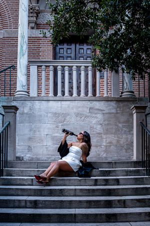 Woman in white dress drinking from bottle on ornate stone steps of historic brick building with balustrade.