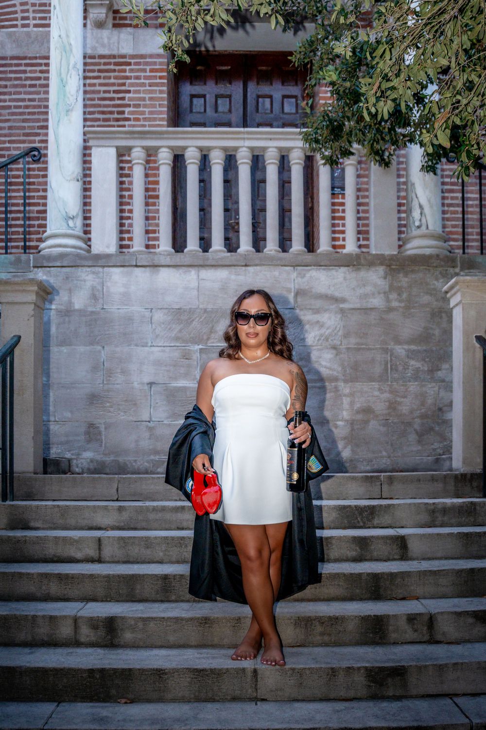 Woman in white strapless dress holding wine bottle on stone steps of brick building.