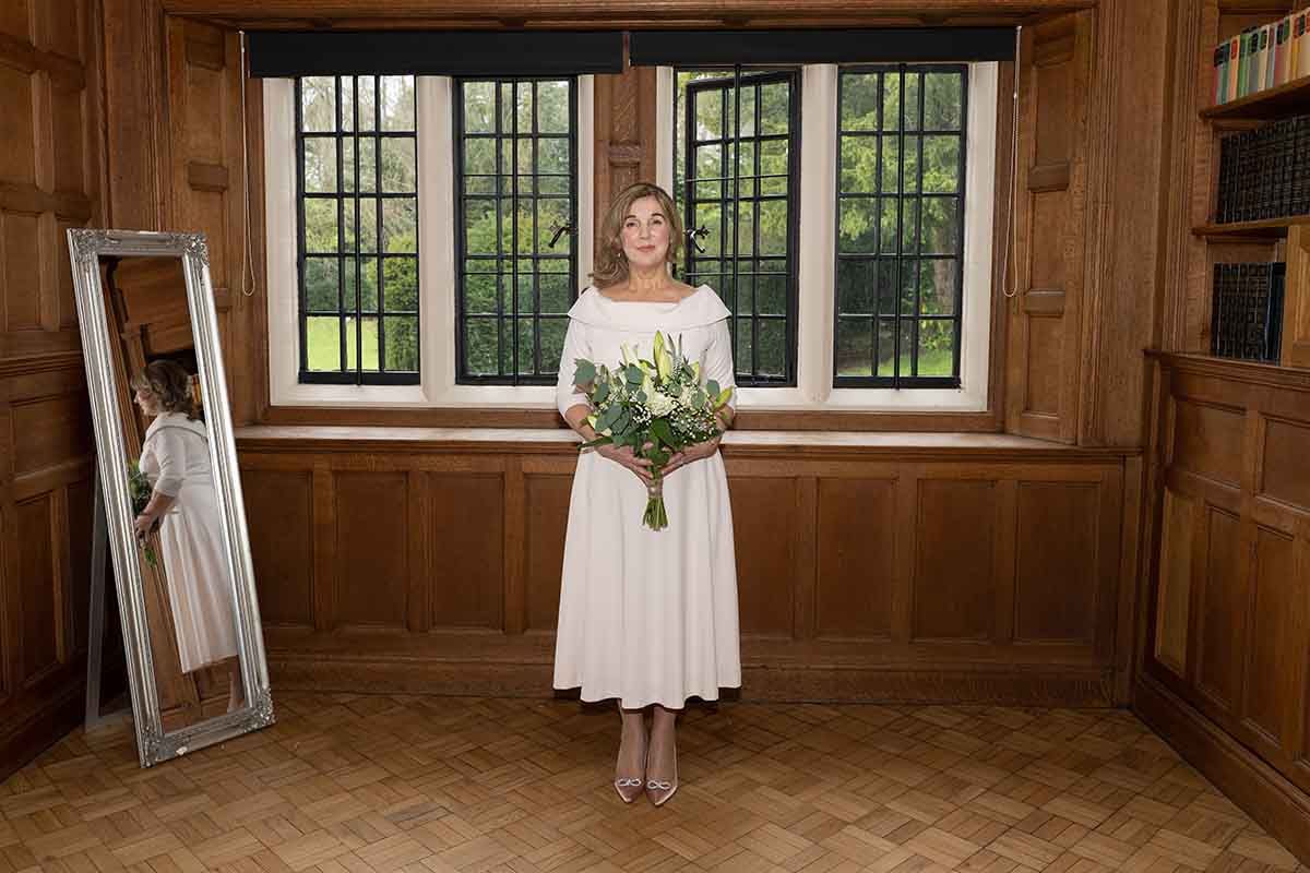 Bride in white midi dress holding bouquet stands in wood-paneled room with mirror reflection and garden window backdrop.