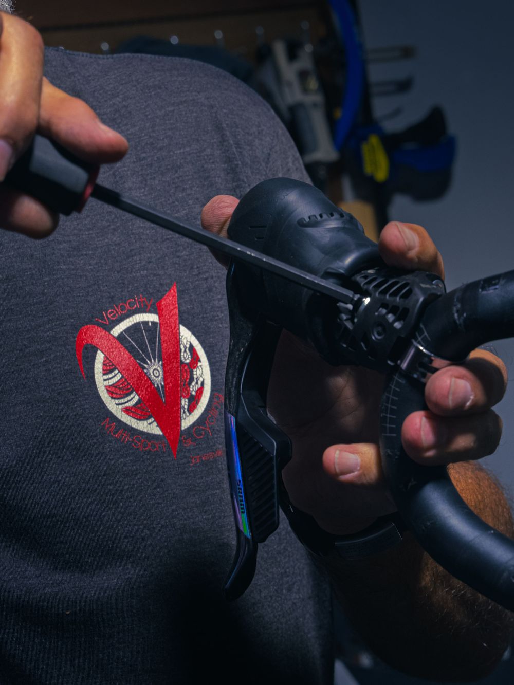 Hands adjusting a SRAM brake lever with a hex wrench, the Velocity Multi Sport & Cycling logo on the mechanic's shirt.