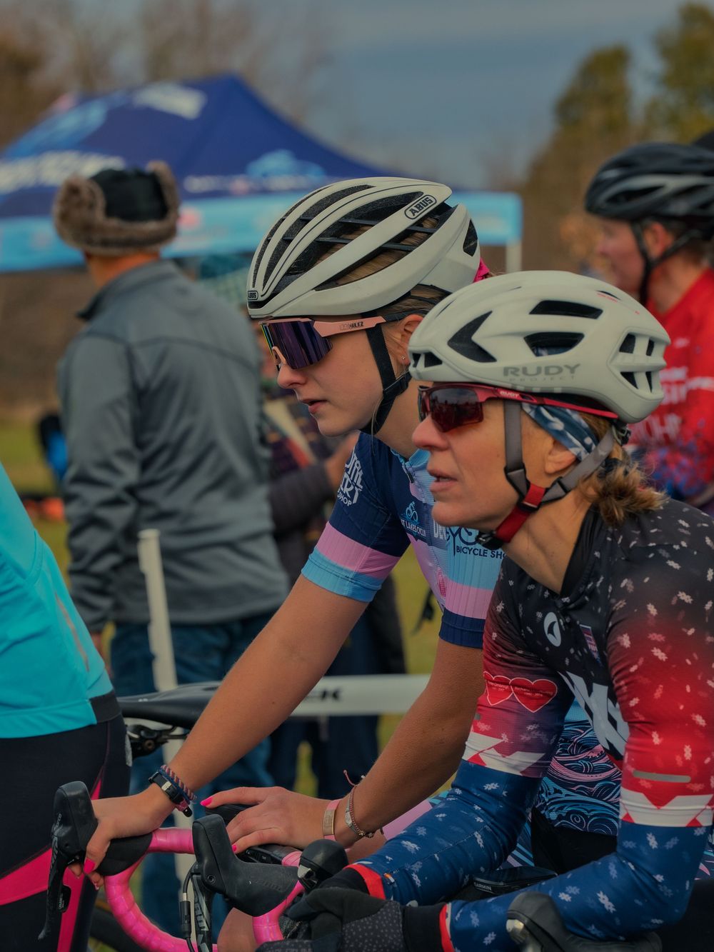 Two cyclists in helmets and sport sunglasses wait side by side at the start line of a cyclocross race, staring ahead as the field lines up.