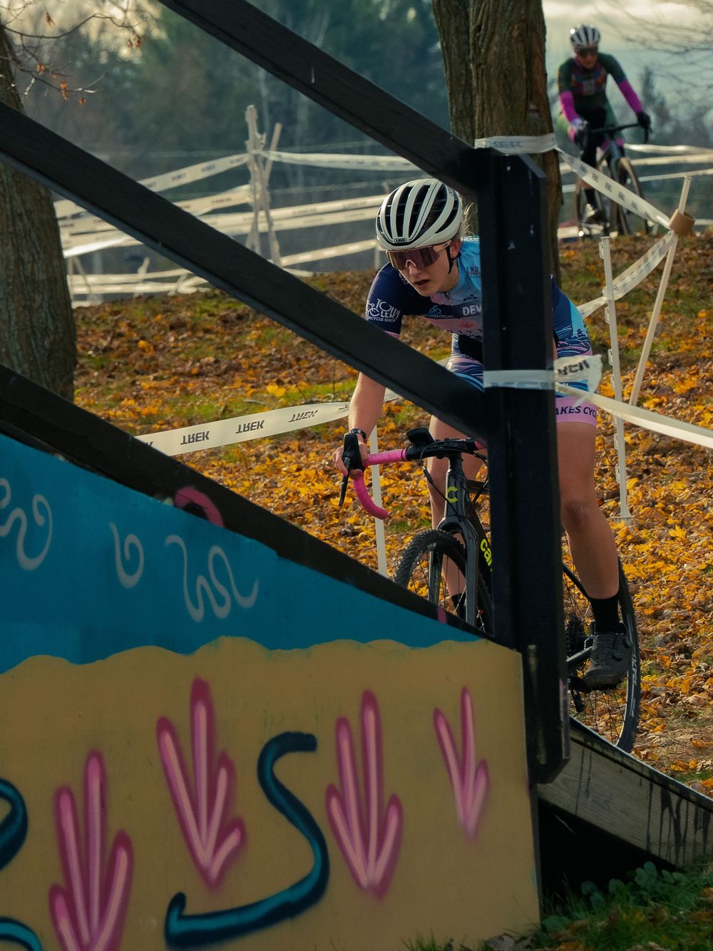 A cyclocross racer rides a wooden ramp past a painted mural at the Trek CX Cup, a second rider chasing through fallen autumn leaves behind.