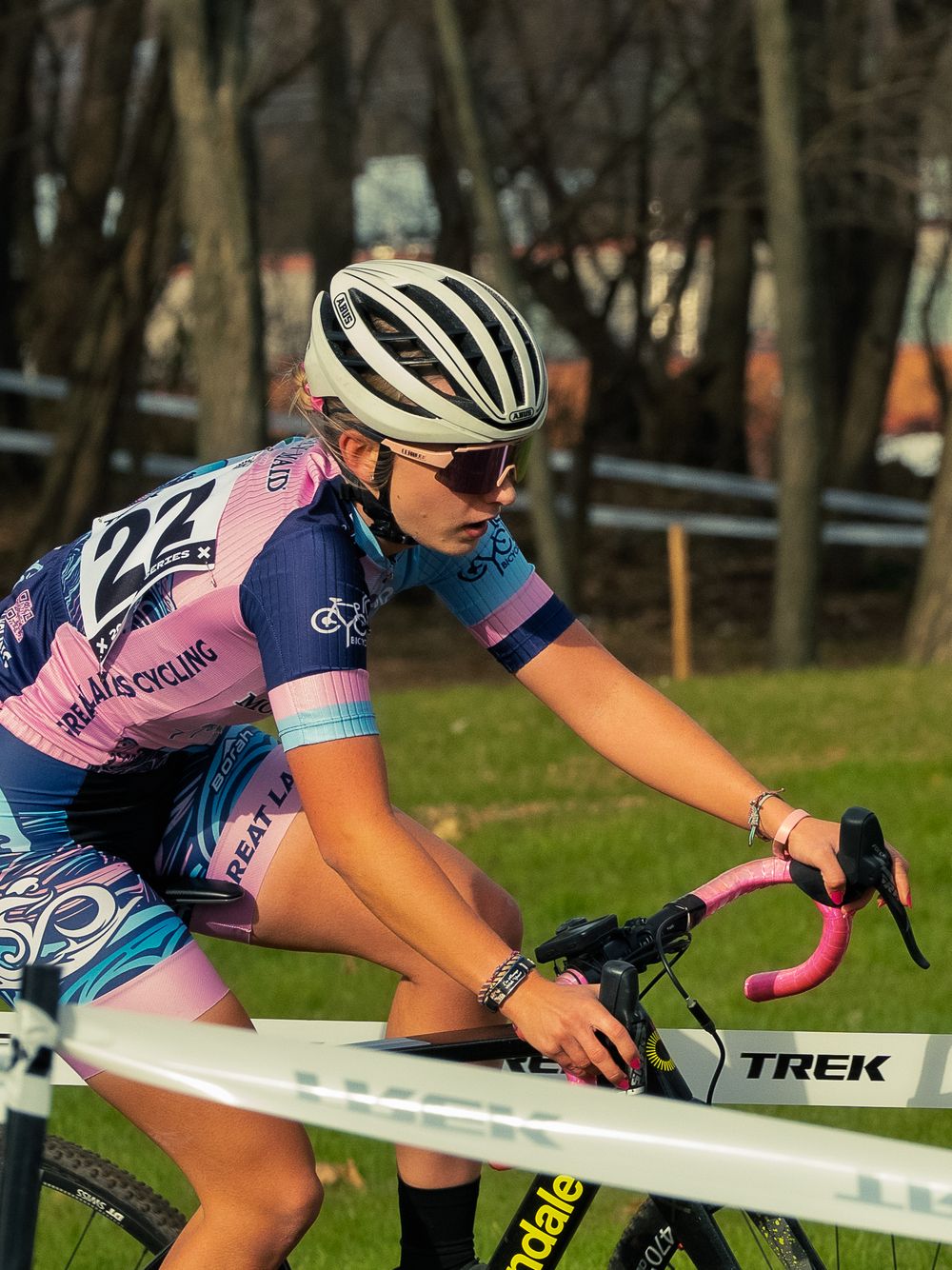 A cyclocross racer in a pink-and-blue kit rides low over pink handlebars past TREK course tape, focused hard on the trail ahead of her.