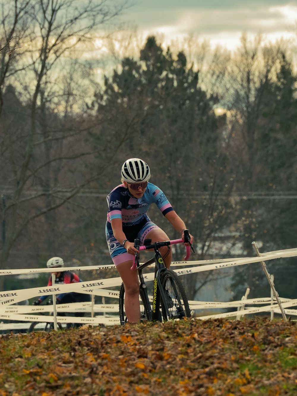 A cyclocross racer crests a leaf-covered rise on a TREK-taped course, a second rider chasing behind under bare autumn trees at dusk.