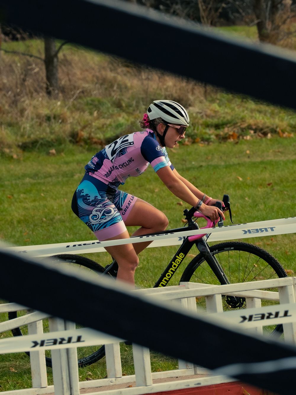 A cyclocross racer in a pink-and-blue kit rides across a wooden bridge on a TREK-taped course, framed through the tape lines from the side.