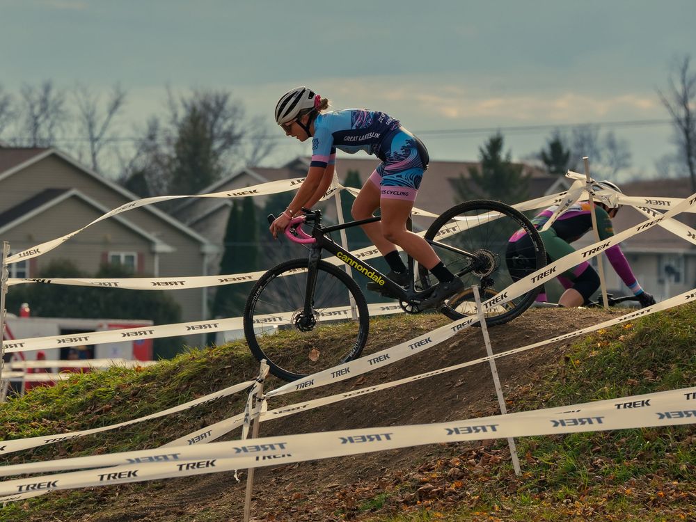 A cyclocross racer in a blue kit rides down a steep dirt bank lined with TREK tape, a chasing rider close behind under a gray autumn sky.