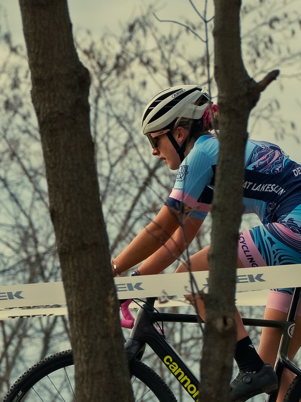 A cyclocross racer in a blue-and-pink kit rides in profile past a TREK-taped course, framed between bare tree trunks under a pale winter sky