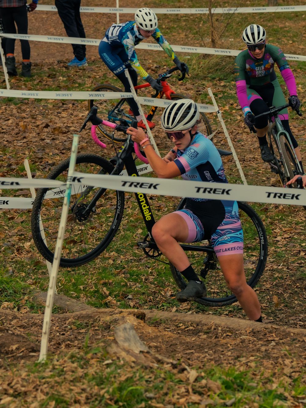 A cyclocross racer shoulders her bike up a leaf-covered climb marked with TREK tape as two other riders push through the course behind her.