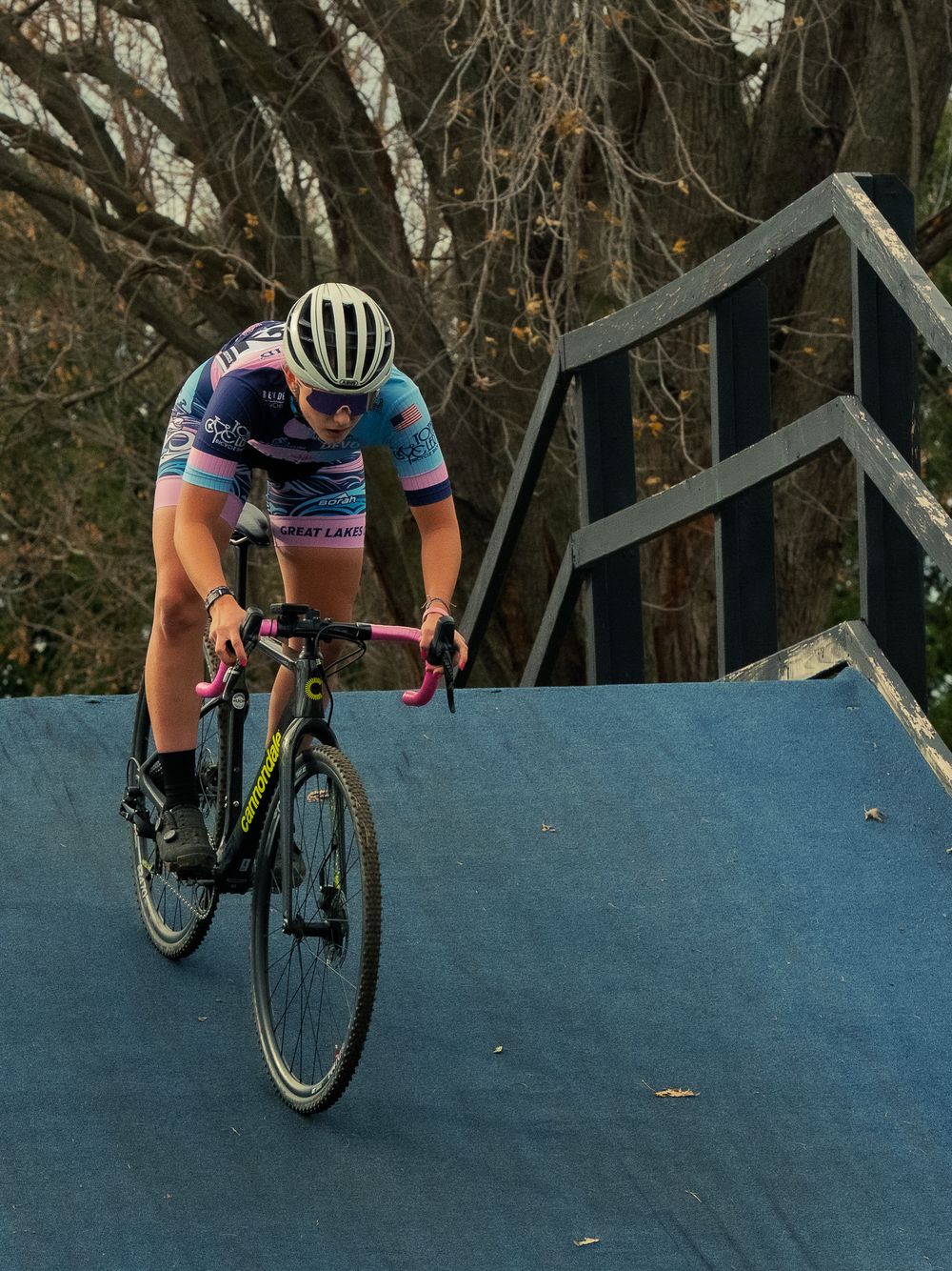 A cyclocross racer in a blue-and-pink kit rides down a steep blue wooden ramp on the course, focused hard with bare autumn trees behind her.