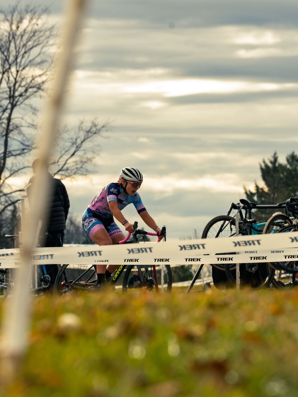 A cyclocross racer in a pink-and-blue kit rides a TREK-taped course past a bike pit at dusk, shot low through blurred grass in foreground.