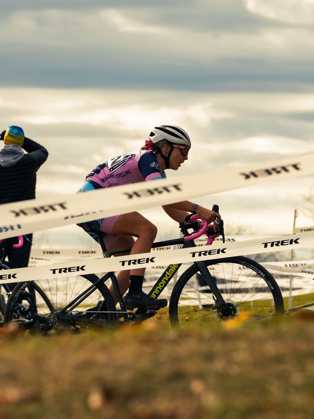 A cyclocross racer in a pink kit rides a course marked with TREK tape under a gray autumn sky, spectators and bikes lining the sideline.