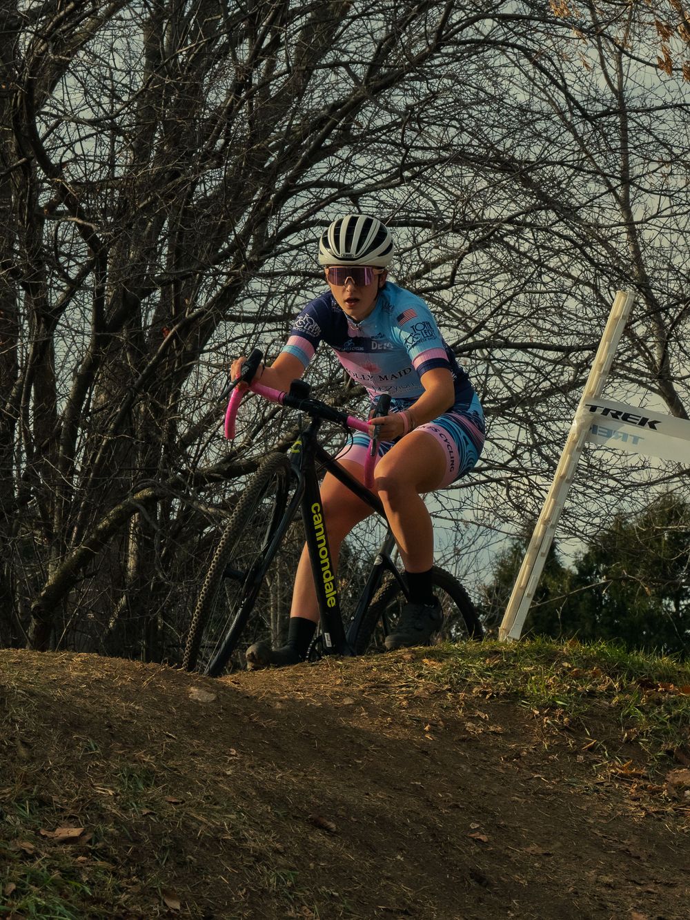 A cyclocross racer in a blue-and-pink kit crests a bare dirt rise on a TREK-taped course, eyes locked ahead under stark leafless trees.