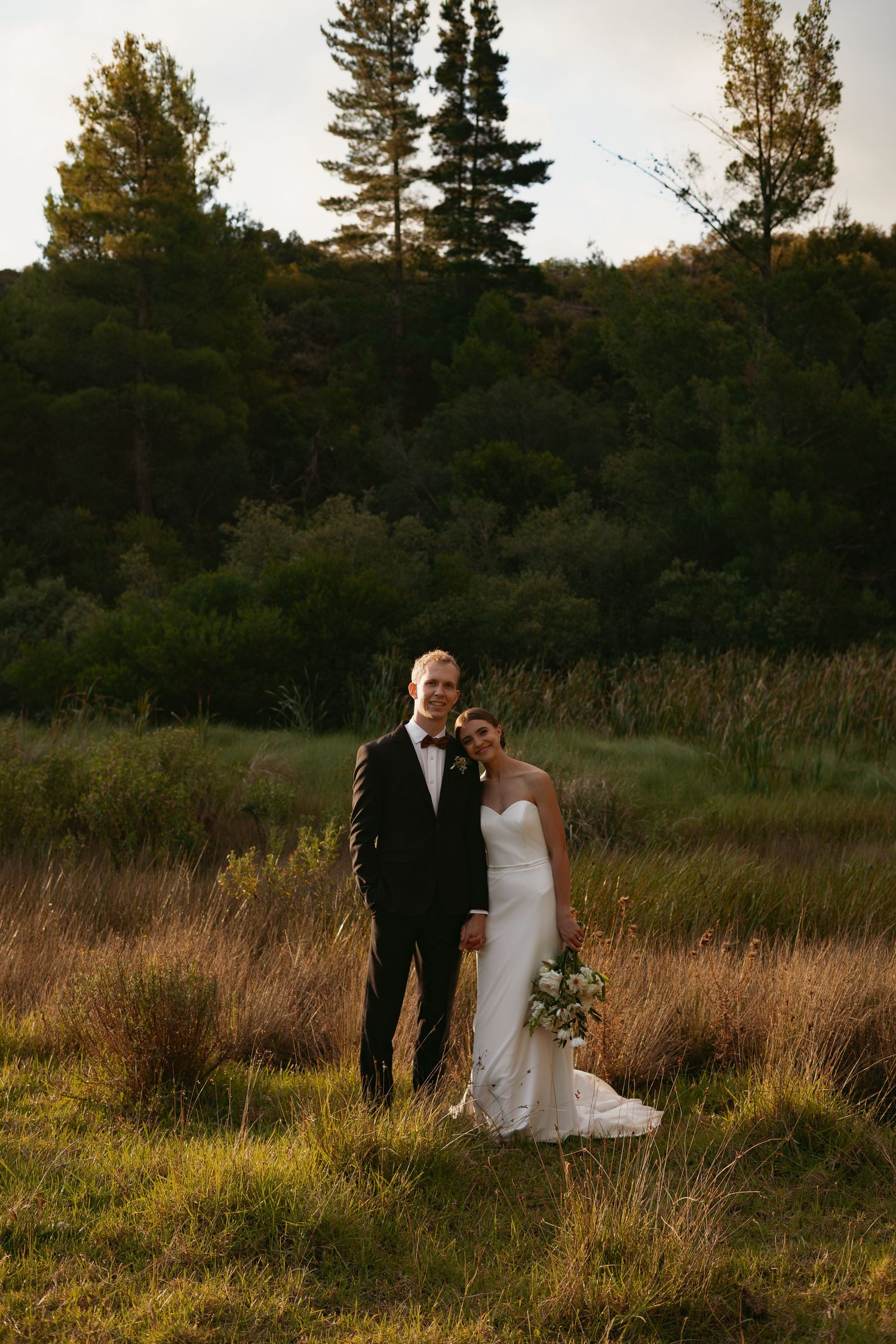 Bride in fitted white gown and groom in black tuxedo standing in golden meadow.