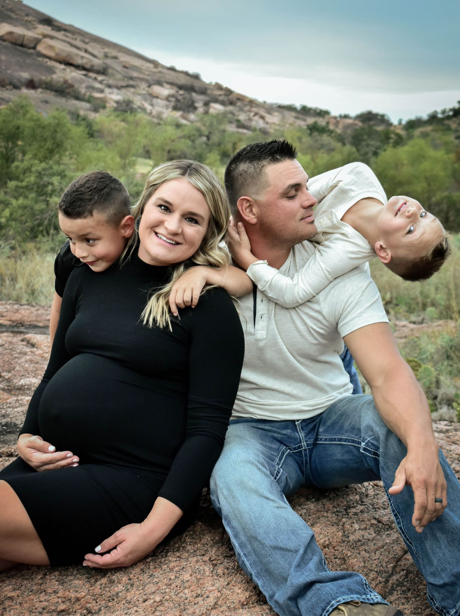 Pregnant woman in black dress poses with husband in white shirt and two young sons on rocky outdoor terrain.