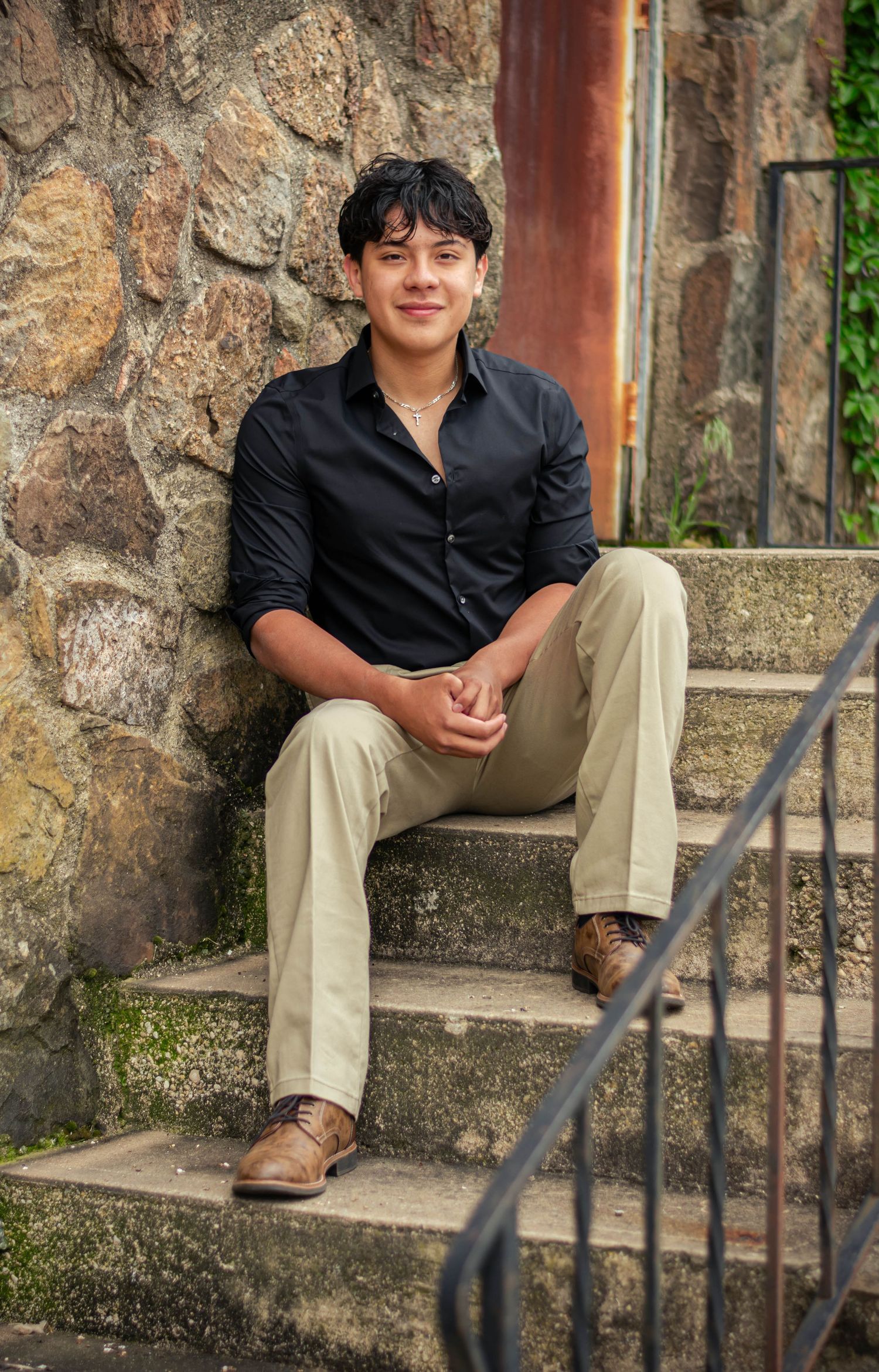 Young man in black shirt and khaki pants sitting on stone steps, smiling confidently in an outdoor portrait.