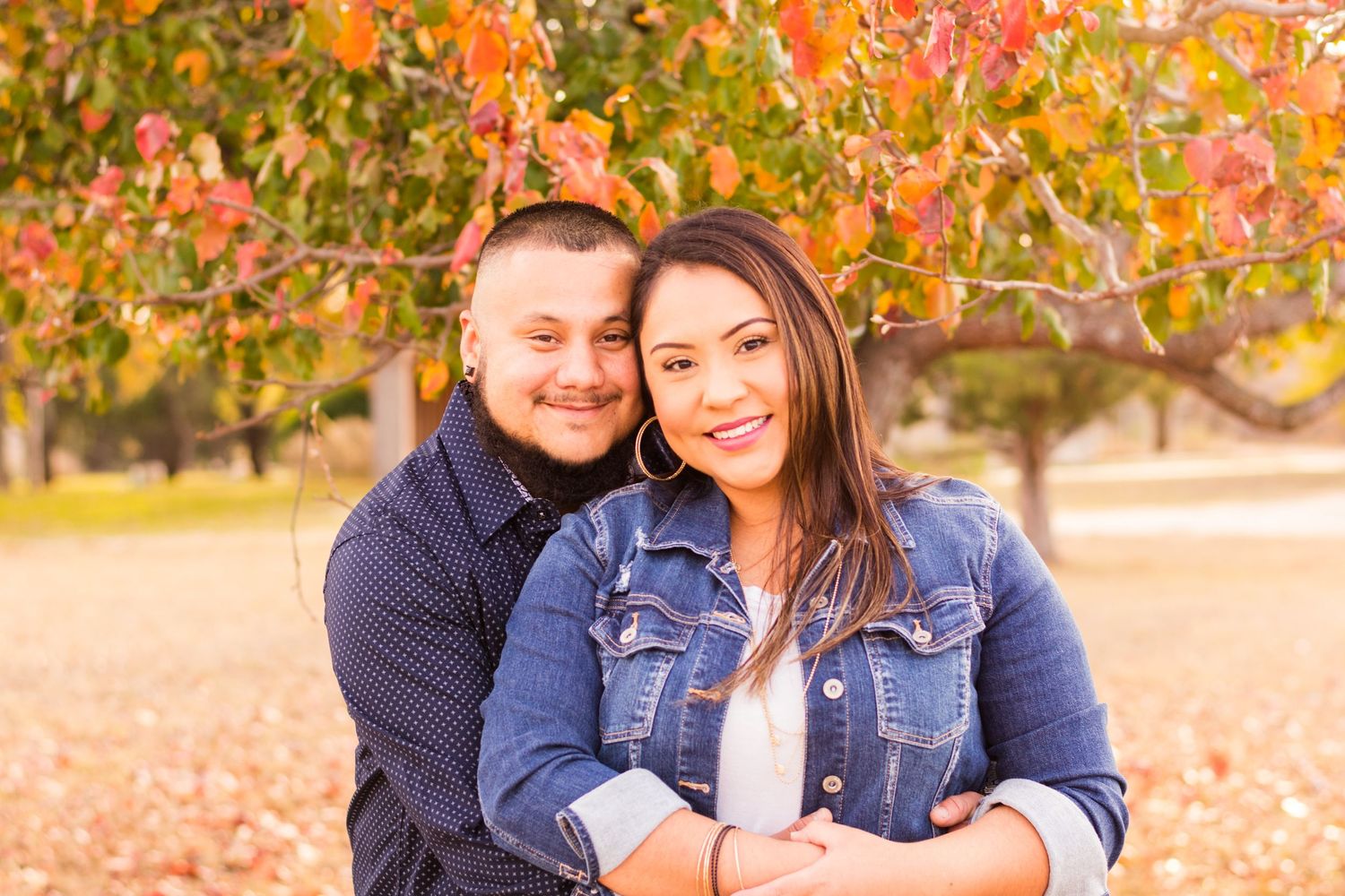 Couple smiling together under colorful autumn foliage, woman in denim jacket, man in navy shirt, fall outdoor portrait.